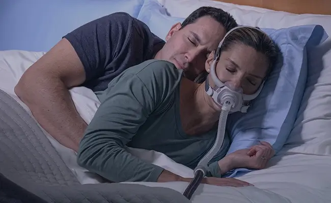 Een koppel dat samen in bed ligt te slapen, waarbij de vrouw een AirFit F40 CPAP-masker draagt.