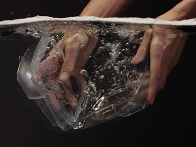 Twee handen dompelen de luchtbevochtigerreservoir van een CPAP-apparaat van ResMed onder in het water, met een zwarte achtergrond
